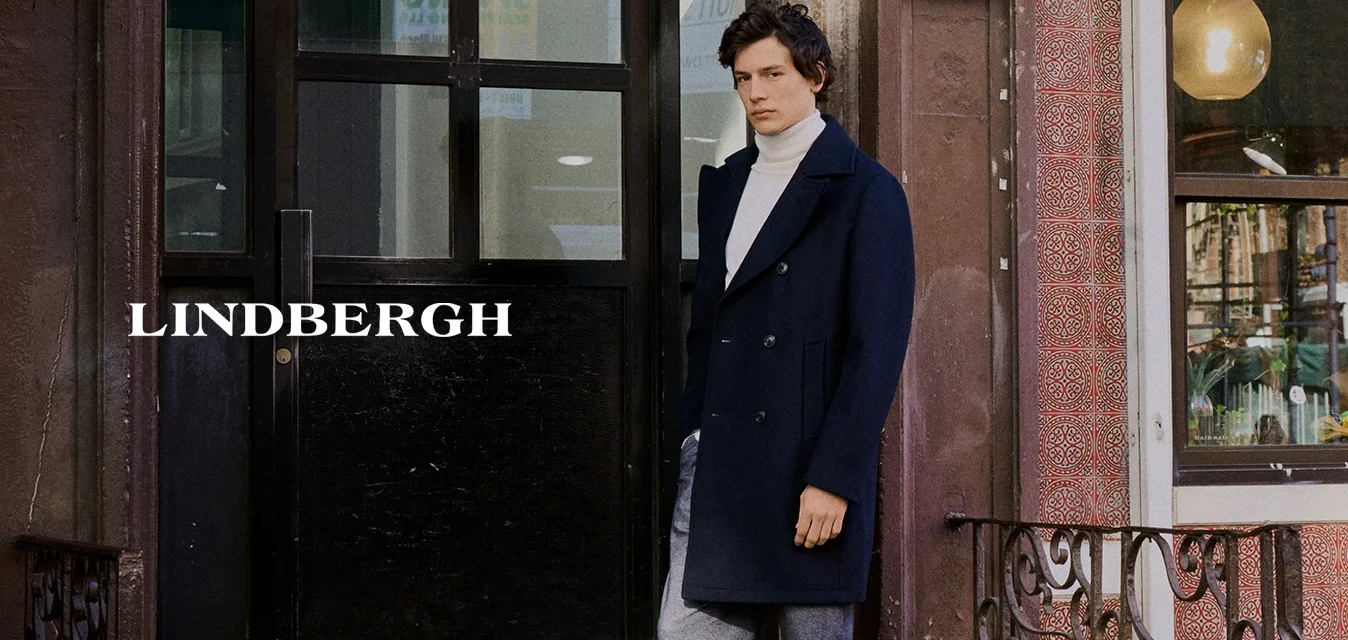 Uomo in cappotto blu marine e pantaloni grigi in piedi su gradini di cemento all'esterno di un edificio in mattoni con grandi finestre e porta nera.