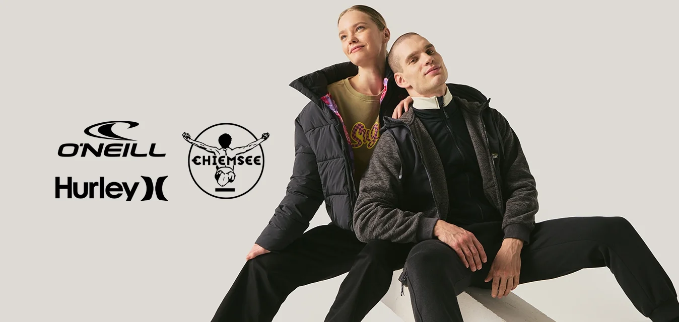 Due persone con abbigliamento casual nero e sneakers posano sedute su blocchi bianchi inclinati contro uno sfondo neutro.