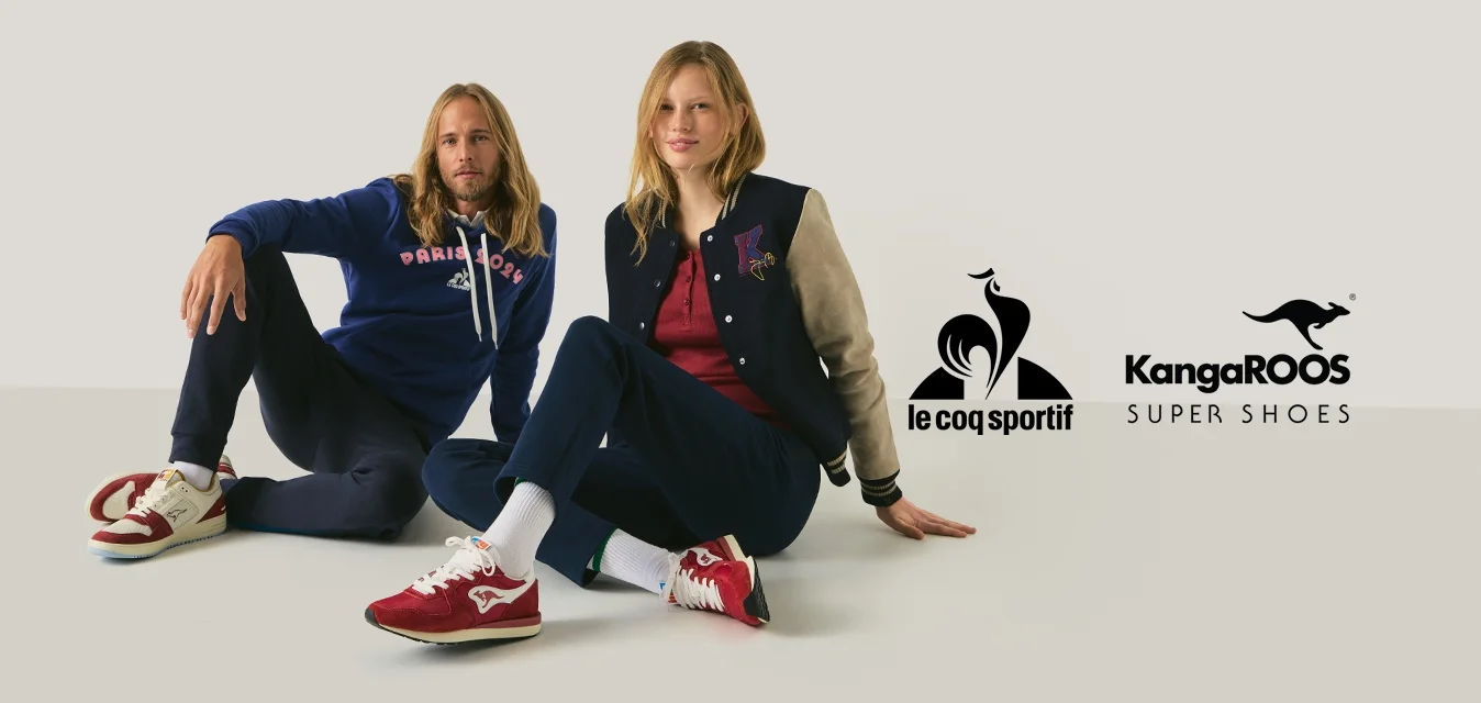 Due persone sono sedute sul pavimento contro uno sfondo semplice, indossano abiti casual e scarpe da ginnastica rosse, appaiono rilassate e guardano verso avanti.