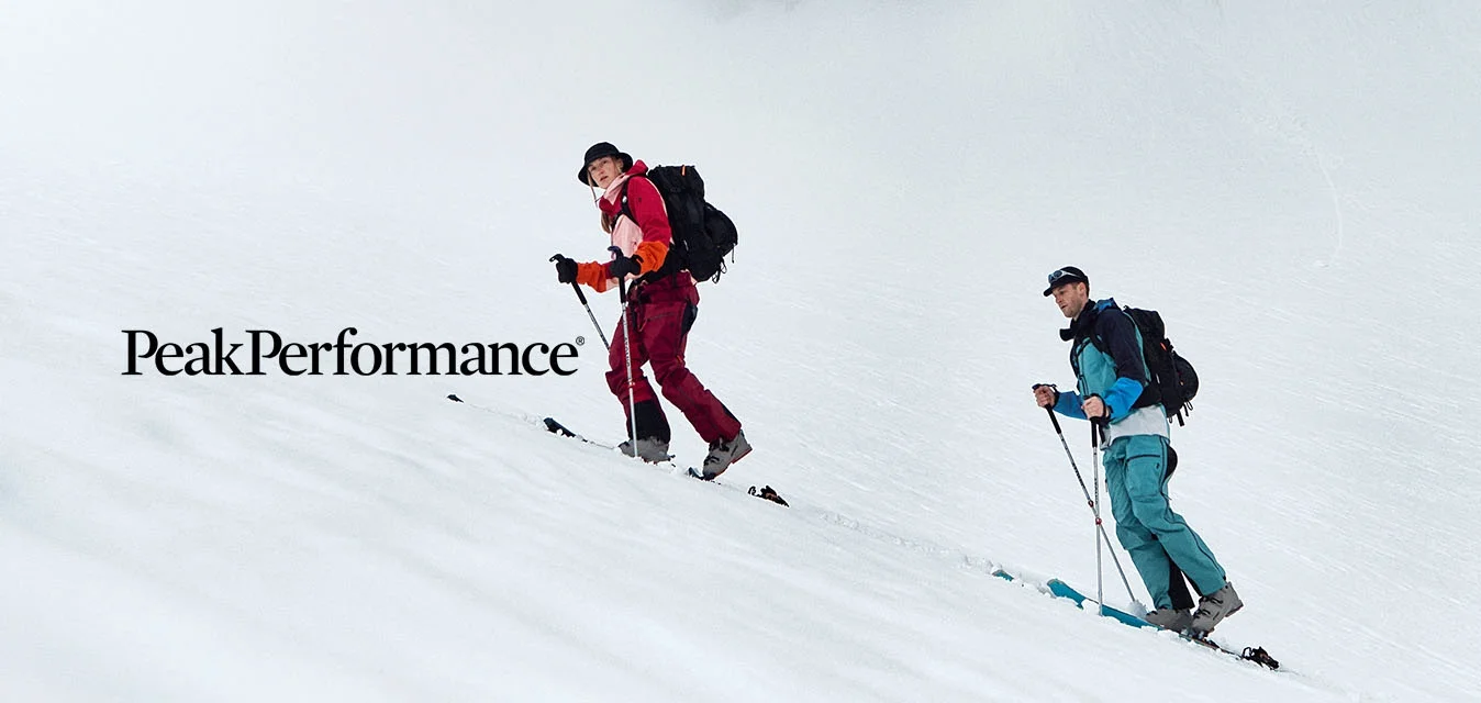 Due escursionisti con abbigliamento invernale e zaini scalano un pendio montano innevato con nebbia e scogliere rocciose sullo sfondo.