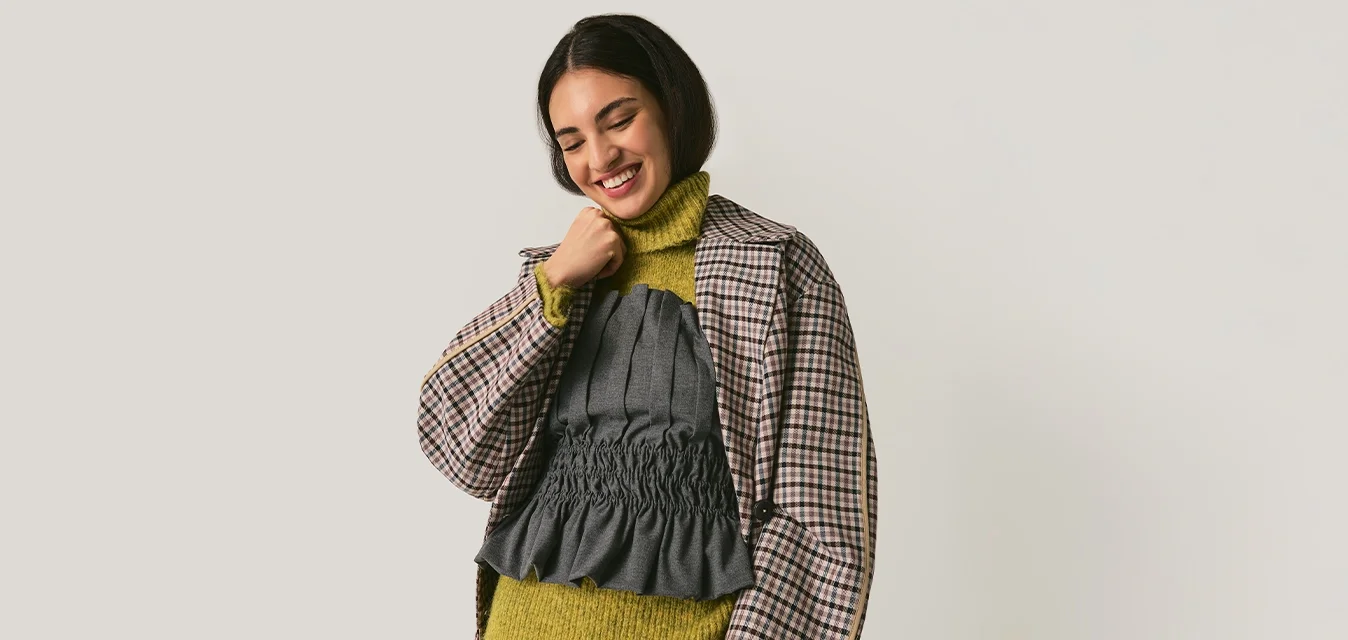 Giovane donna sorridente, indossa un blazer a quadri sopra un maglione verde a collo alto e una maglia grigia con volant, con pantaloni grigi a righe, in piedi davanti a uno sfondo neutro.