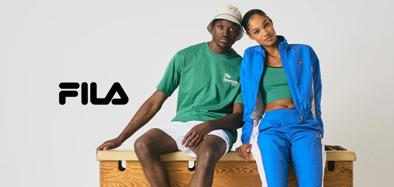 Due giovani adulti in abbigliamento sportivo casual seduti su una cassa da palestra di legno contro uno sfondo grigio semplice, rilassati e inclinati l'uno verso l'altro.