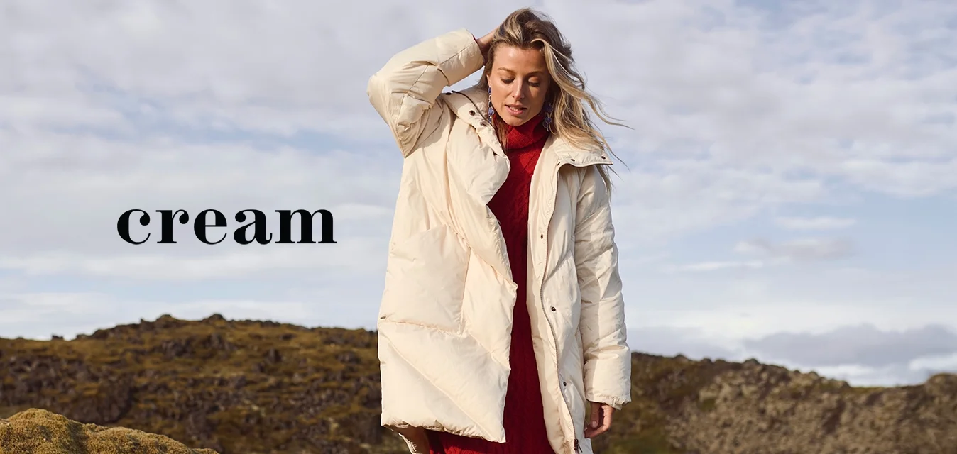 Cappotto bianco gonfio con grandi tasche, indossato sopra un abito rosso a coste, abbinato a stivali alti color sabbia, su uno sfondo di paesaggio muschioso.