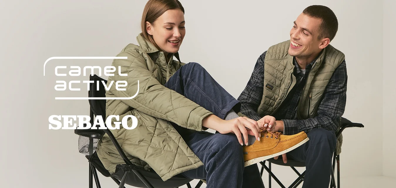 Due persone con indumenti esterni verdi sono sedute su sedie pieghevoli nere; una sistema la scarpa marrone dell'altra, entrambe sorridono in una stanza bianca minimalista.