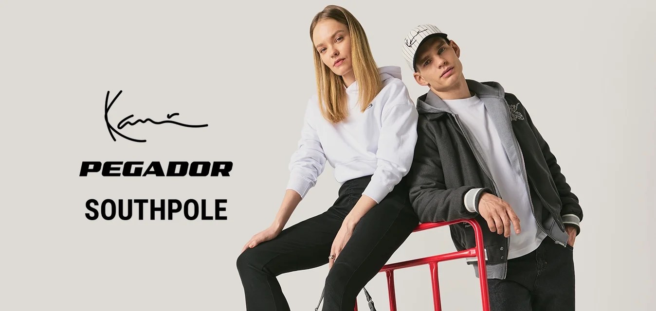 Una donna con felpa bianca con cappuccio è seduta su un carrello rosso a piattaforma mentre un uomo con giacca nera e cappellino si appoggia al manico in un ambiente da studio.