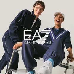 Due giovani uomini in tute blu marino sorridenti, uno spinge l'altro seduto su un carrello di metallo su uno sfondo neutro.