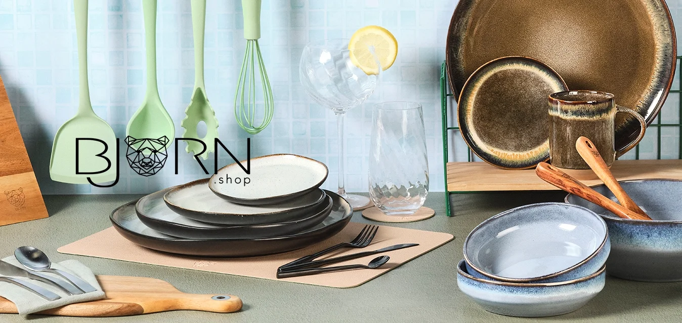 Quattro utensili da cucina verdi appesi, coltelli in ceppo di legno, piatti e ciotole in ceramica impilati, un bicchiere con fetta di limone e cucchiai di legno.