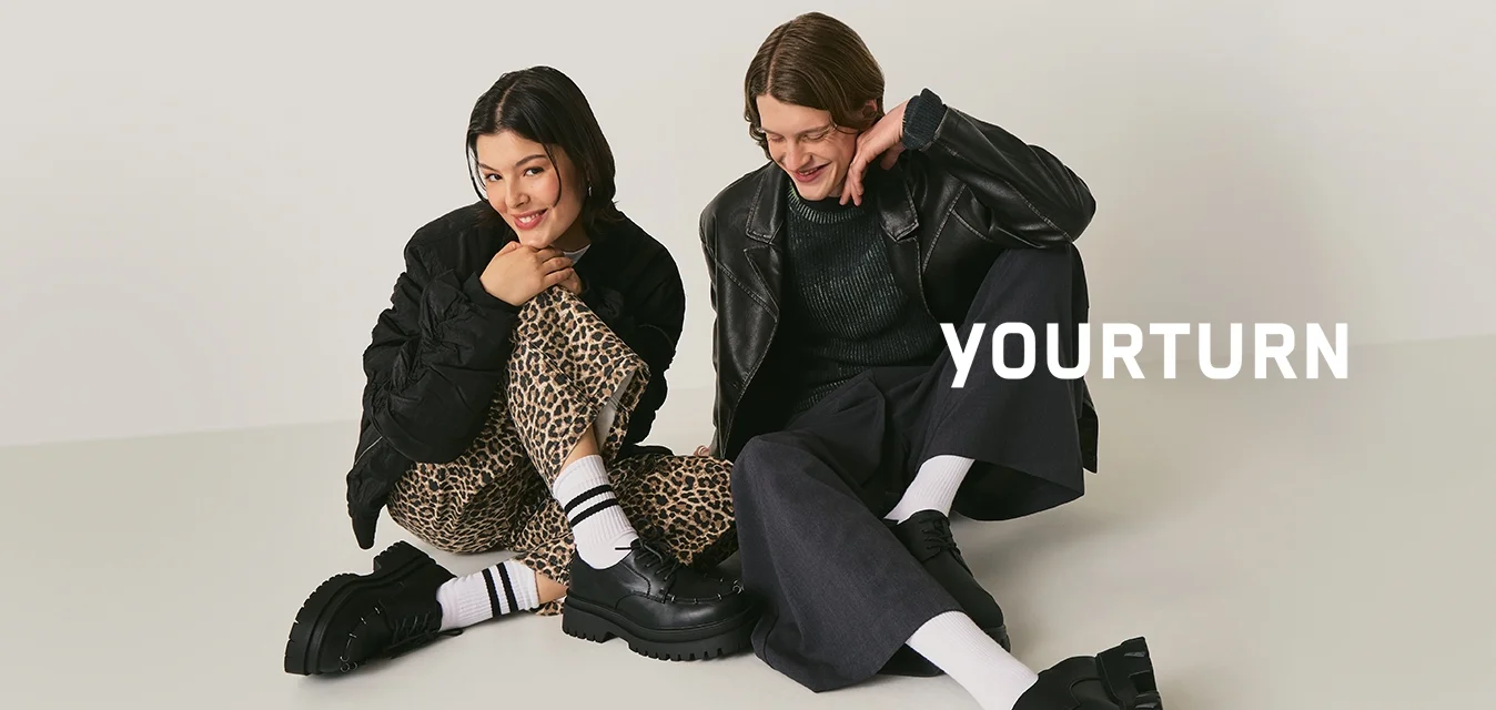 Due persone sedute per terra indossano giacche nere, calze bianche e scarpe nere chunky; una indossa pantaloni con stampa leopardata, l'altra dei pantaloni larghi scuri.