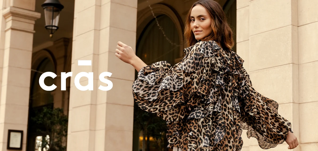 Donna che indossa una camicetta trasparente con stampa leopardata e pantaloni grigio chiaro, in piedi sotto archi di pietra, si volta con un leggero sorriso.