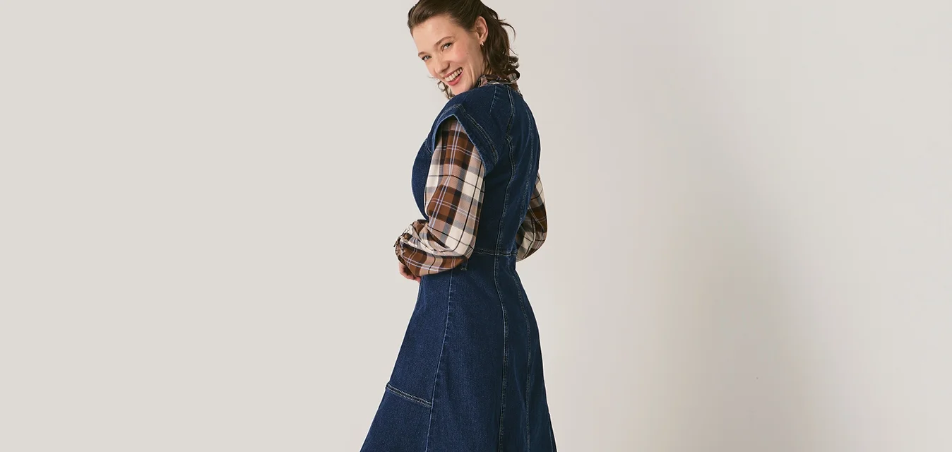 Donna in un lungo abito di denim blu scuro sopra una camicia a quadri, con stivaletti marroni, in piedi e che si volta sorridendo su uno sfondo chiaro e semplice.