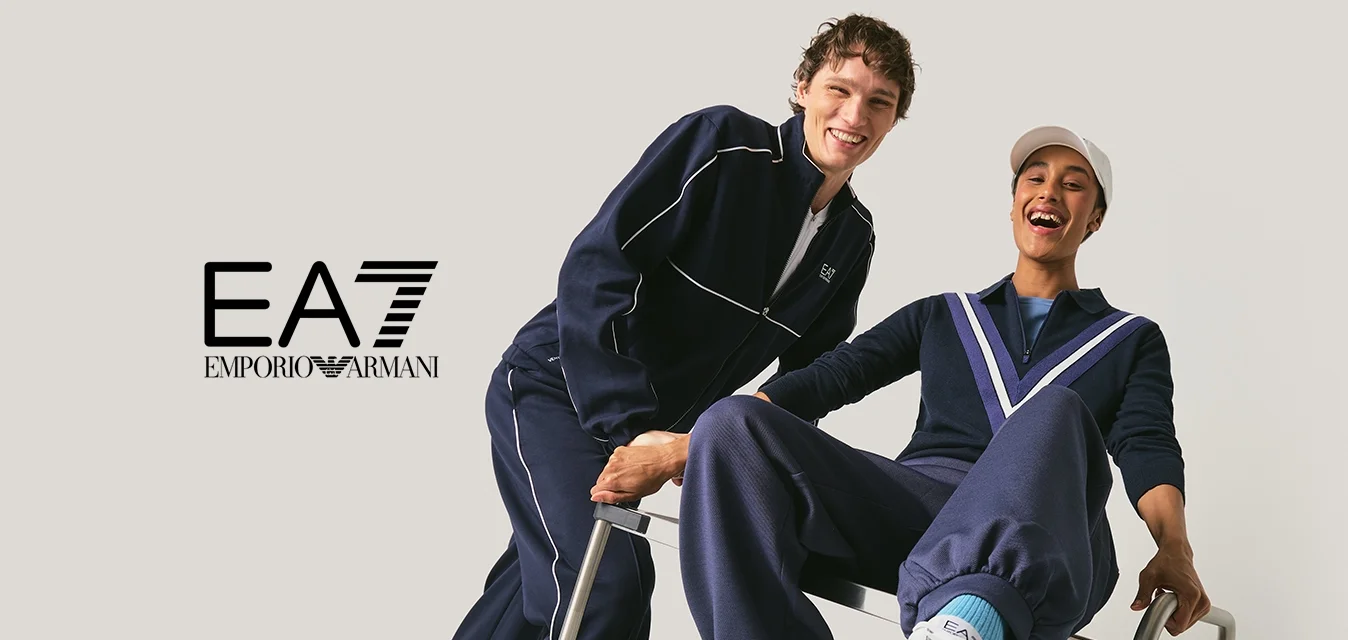 Due giovani uomini in tute blu marino sorridenti, uno spinge l'altro seduto su un carrello di metallo su uno sfondo neutro.