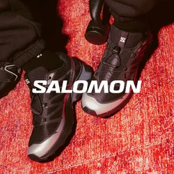 Scarpe da ginnastica nere con dettagli grigi, tomaia in mesh testurizzato e suola in gomma sono appoggiate su un tappeto rosso, con dei pantaloni della felpa neri larghi in vista.