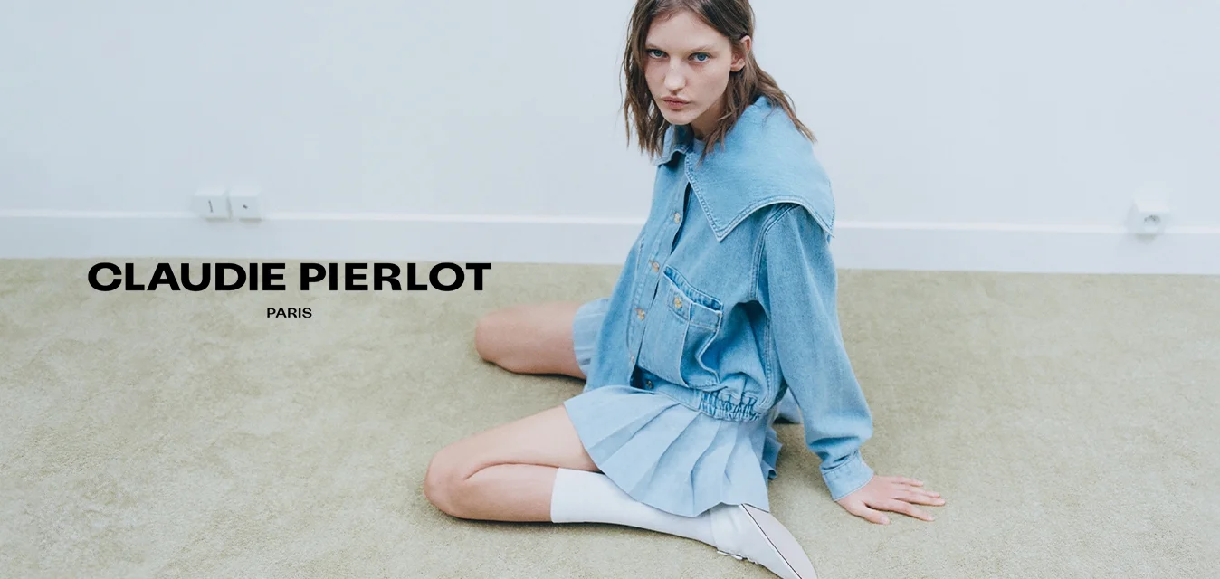 Giovane donna con capelli bagnati che indossa una giacca di jeans oversize e una gonna plissettata, seduta su un pavimento tappezzato contro una parete bianca uniforme.