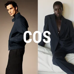 Uomo con giacca blu scuro e pantaloni neri in piedi di lato; donna con completo grande e scuro seduta su un divano bianco contro una parete liscia.