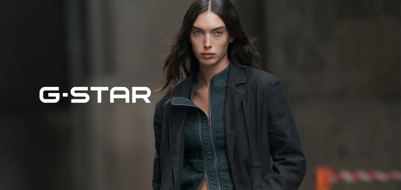 Donna con capelli lunghi e scuri che indossa una giacca scura aperta sopra un denim aderente e un blazer scuro, in piedi in un ambiente urbano.