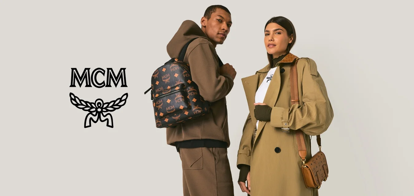 Uomo con felpa con cappuccio marrone e pantaloni con zaino a motivi; donna con cappotto lungo beige e maglietta bianca che tiene una borsa a tracolla marrone, in piedi.