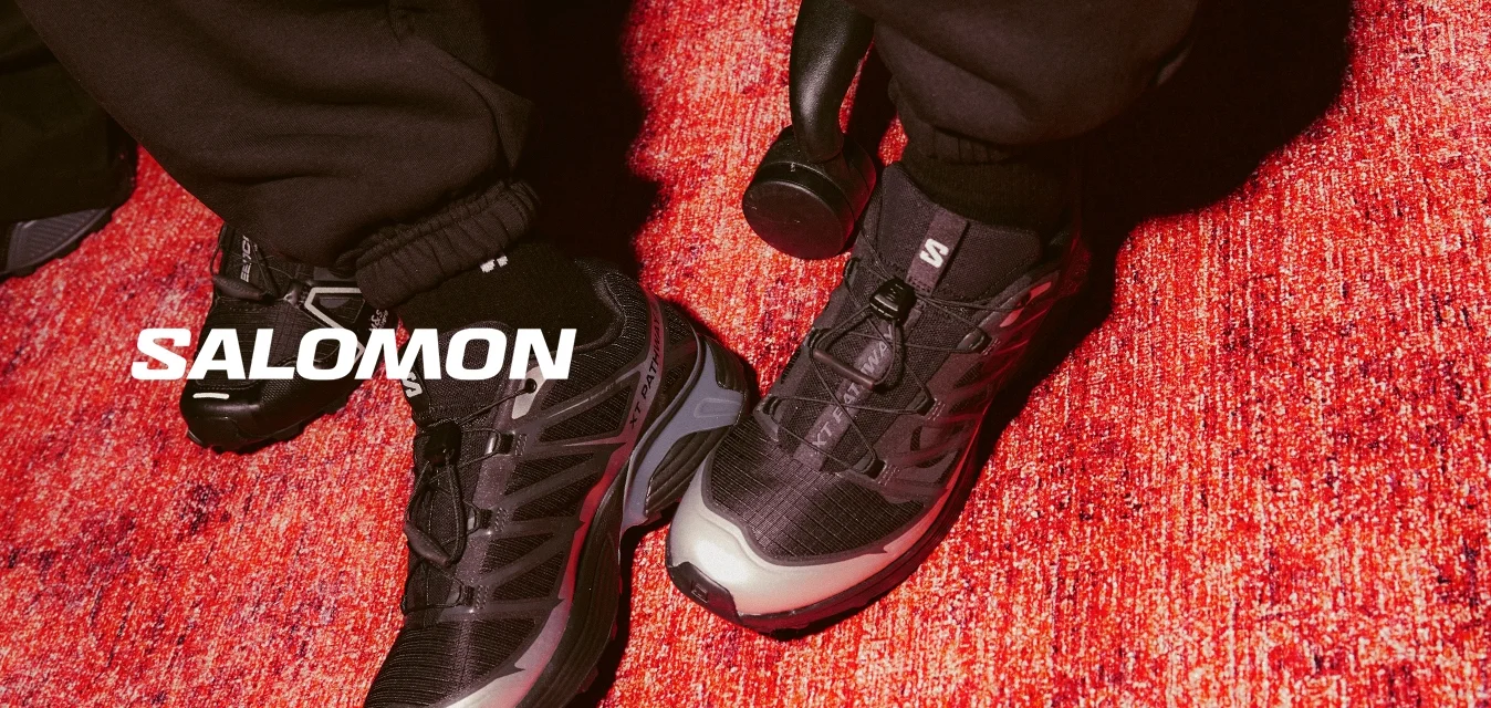 Scarpe da ginnastica nere con dettagli grigi, tomaia in mesh testurizzato e suola in gomma sono appoggiate su un tappeto rosso, con dei pantaloni della felpa neri larghi in vista.