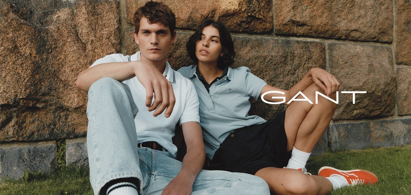 Due persone sono sedute contro un muro di pietra. L'uomo indossa una polo bianca e jeans chiari; la donna indossa una polo azzurra e pantaloni corti neri.