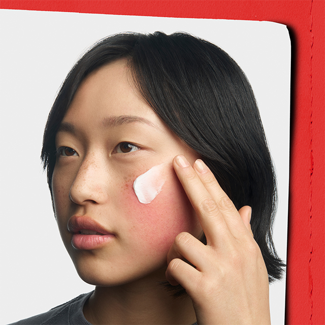 Donna che applica una crema viso sulla guancia arrossata con le dita; il rossore &egrave; visibile nell&rsquo;area di applicazione.