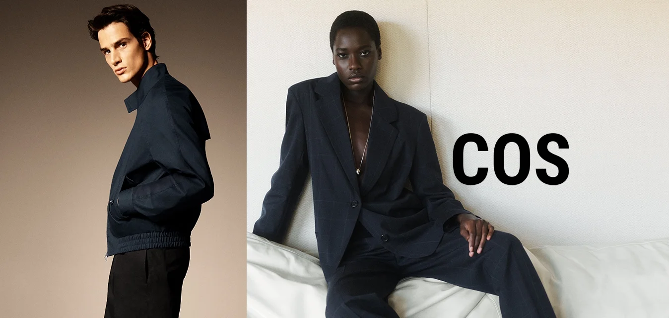 Uomo con giacca blu scuro e pantaloni neri in piedi di lato; donna con completo grande e scuro seduta su un divano bianco contro una parete liscia.