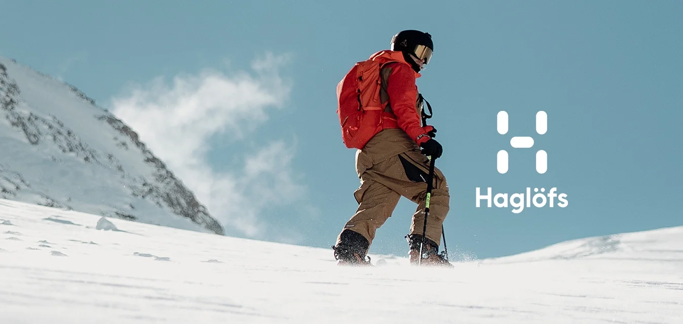 Persona con giacca rossa e berretto nero che cammina su una pendenza innevata con uno sfondo montano. Cielo azzurro limpido e superficie della neve texturizzata visibili.