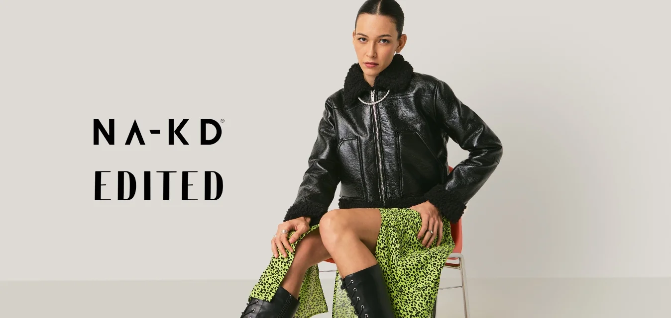 Donna seduta su una sedia, indossa una giacca di pelle nera, una gonna divisa verde neon e stivali alti neri con lacci, su sfondo neutro.