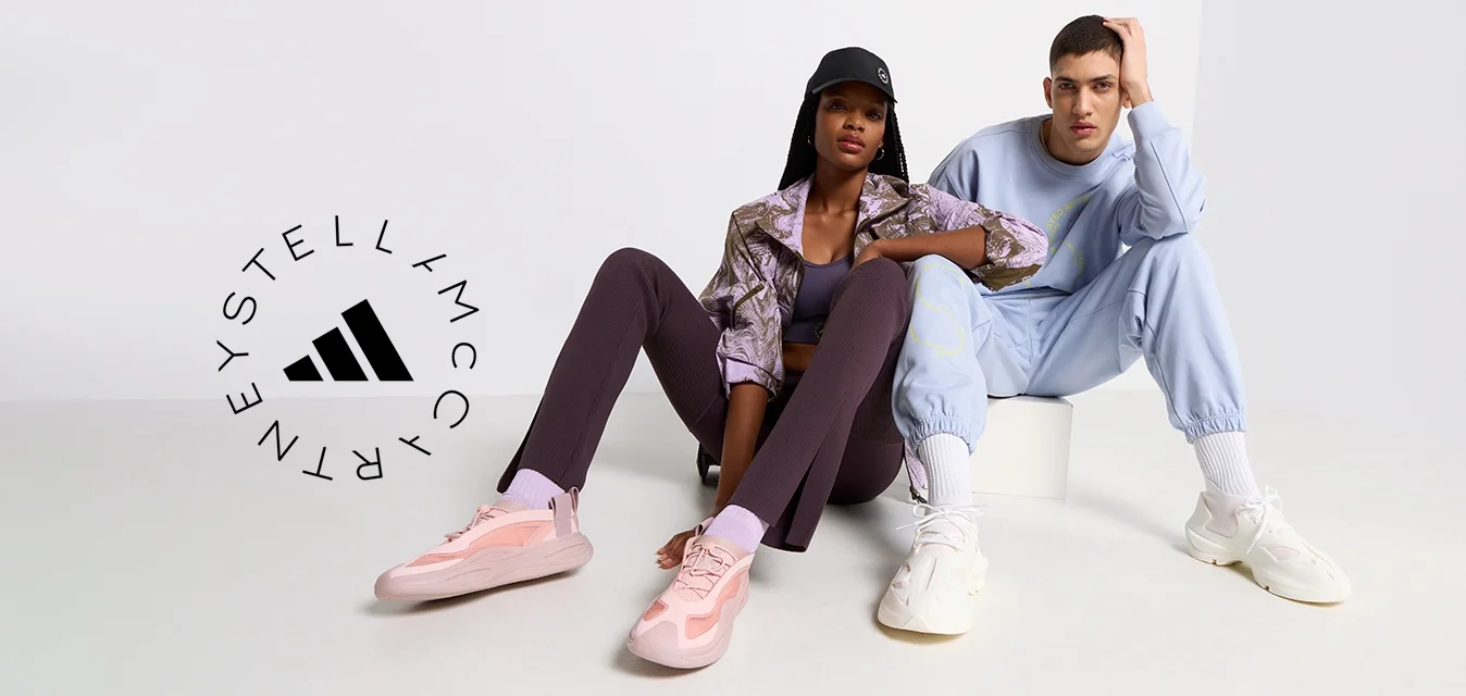 Due modelli seduti su una superficie chiara. Uno indossa un giacca marrone a fantasia, leggings viola e scarpe da ginnastica rosa. L'altro indossa un tuta blu chiaro e scarpe da ginnastica bianche.