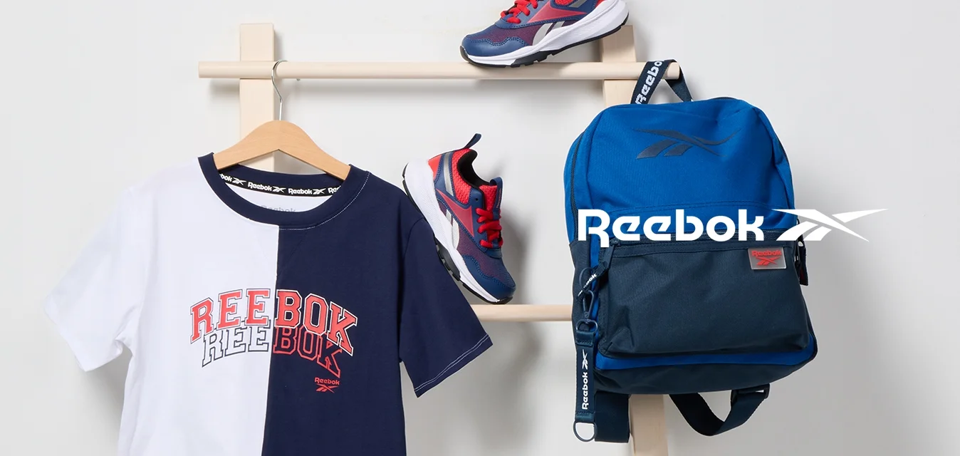 T-shirt Reebok bicolore appeso a un gancio, un paio di sneaker rosse e blu navy su una scala di legno, e uno zaino blu Reebok appeso.