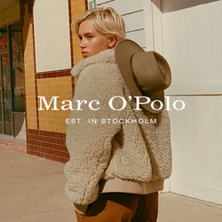 Giacca in shearling beige e pelosa con colletto alto, indossata sopra un maglione crema. Pantaloni marroni, cappello a tesa larga verde appoggiato sulla giacca.