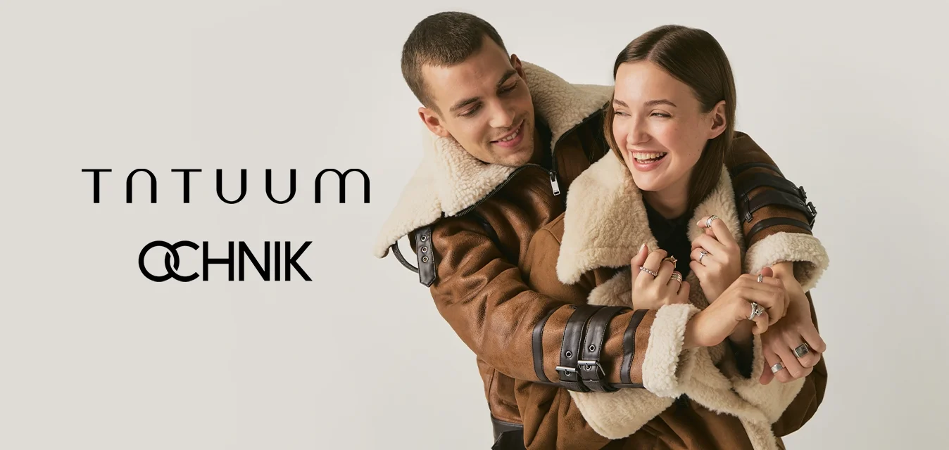 Giovane coppia sorridente che si abbraccia, entrambi indossano giacche di shearling marrone abbinate su uno sfondo chiaro e semplice.