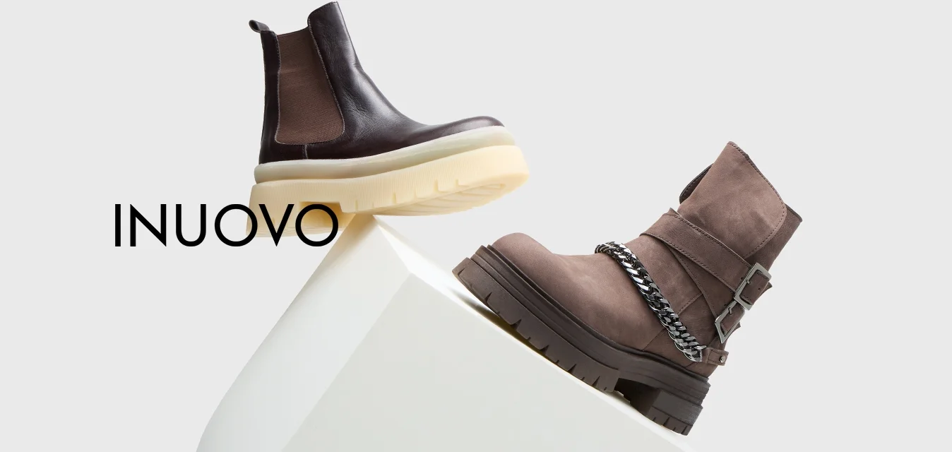 Due stivaletti: uno in pelle marrone con una suola spessa beige, l'altro in suede grigio con un accento a catena e suola in gomma nera, esposti su un piedistallo bianco.