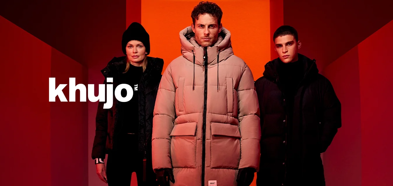 Tre persone con cappotti invernali e stivali si trovano in un corridoio illuminato di rosso con pareti e pavimento geometrici.