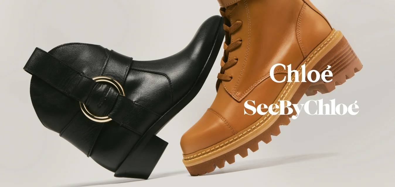 Stivaletto nero alla caviglia con dettaglio ad anello dorato di fronte a uno stivale da combattimento color cuoio con lacci e doppie fibbie su sfondo beige.