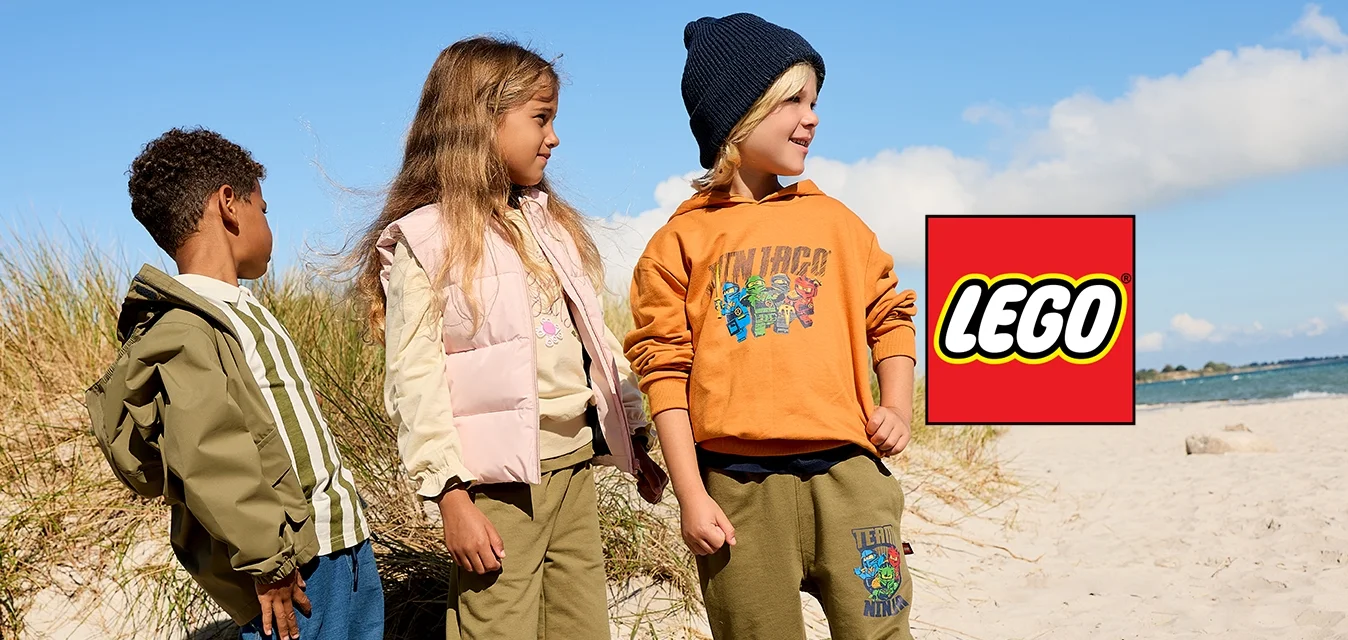 Tre bambini in piedi su una spiaggia sabbiosa vicino alle dune di erba sotto un cielo azzurro limpido, vestiti con abiti autunnali casual.