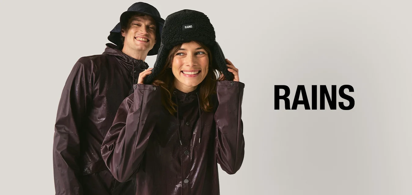 Due persone con giacche antipioggia viola scuro e cappelli neri sorridono davanti a uno sfondo chiaro e uniforme.