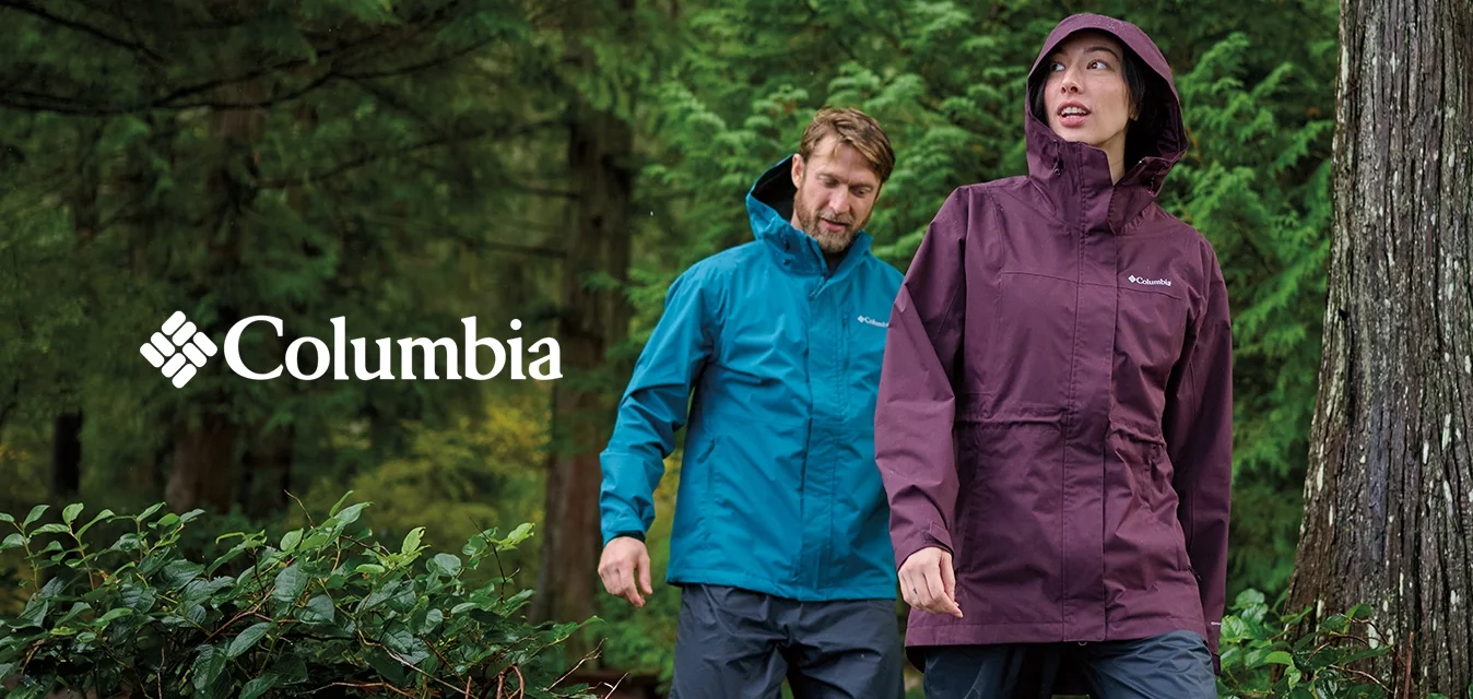 Due escursionisti con giacche antipioggia blu e viola camminano attraverso una fitta foresta verde con grandi alberi e arbusti.