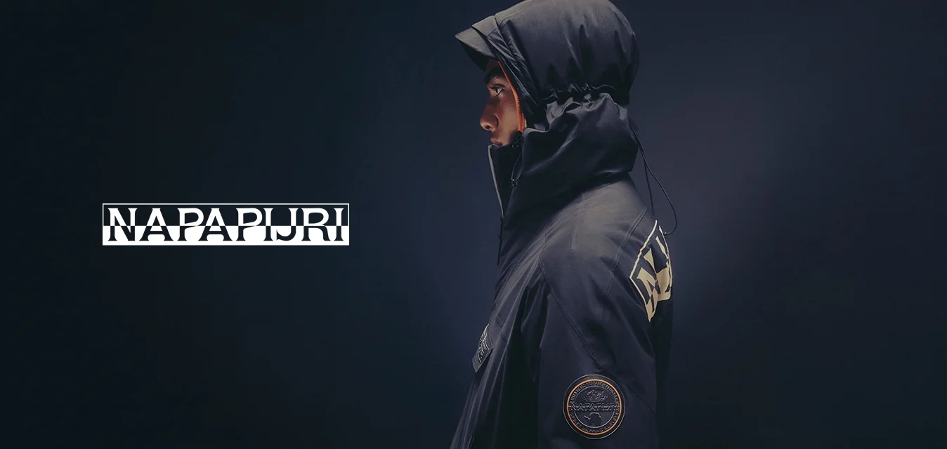 Persona di profilo che indossa una giacca scura con cappuccio e un cappello a tesa, con una toppa rotonda sulla manica e un grande testo sulla schiena.