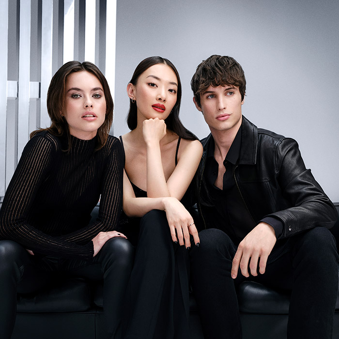 Tre modelli in eleganti abiti neri posano insieme su uno sfondo grigio moderno.