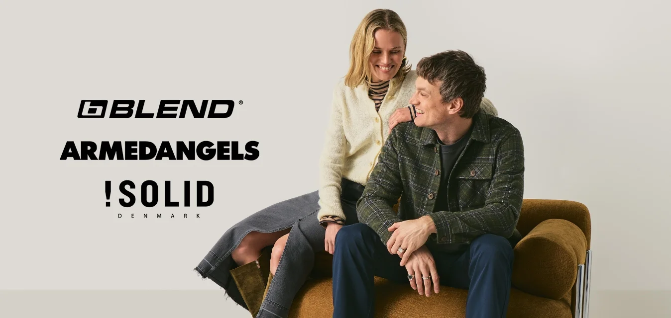 Un uomo e una donna sono seduti vicini su una poltrona imbottita marrone, davanti a uno sfondo beige uniforme, sorridendo e guardandosi a vicenda.