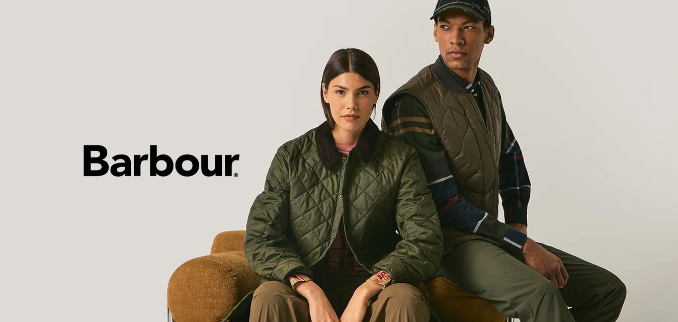 Due persone indossano giacche trapuntate verde oliva sedute su una poltrona marrone testurizzata, su uno sfondo beige uniforme.