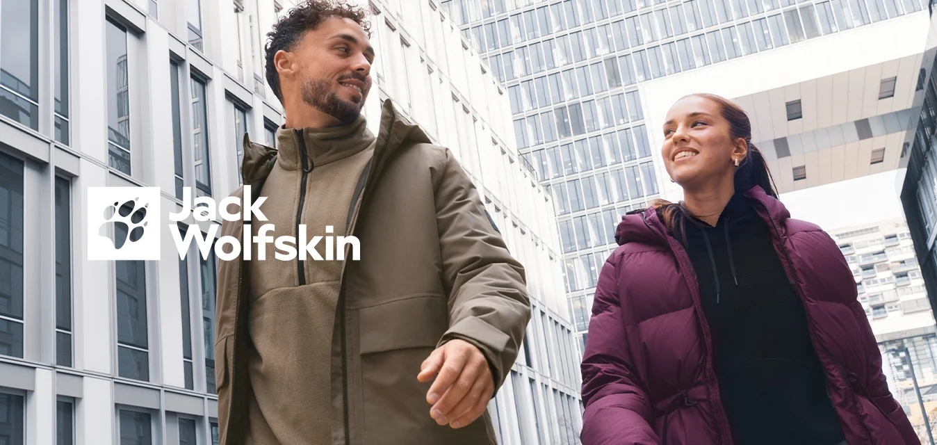 Due persone con giacche invernali camminano e sorridono all'aperto davanti a moderni uffici in un contesto urbano in una giornata nuvolosa.
