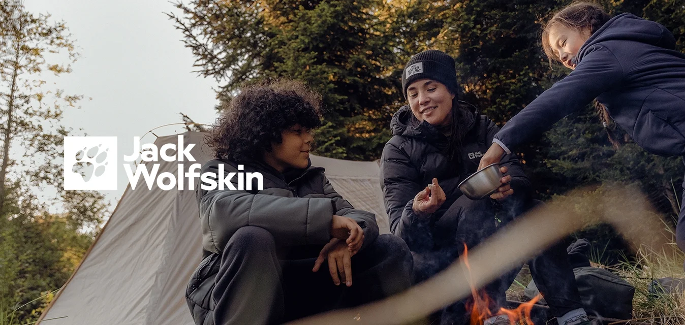 Tre persone siedono attorno a un falò con una tenda beige sullo sfondo. Indossano giacche imbottite e stivali, circondati da erba e alberi.