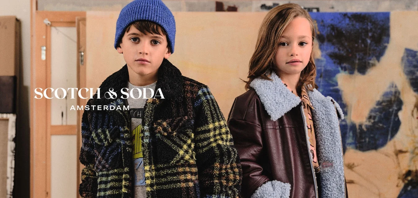 Due bambini stanno in uno studio d'arte. Il ragazzo indossa un cappotto di lana a quadri, una camicia grigia, jeans e stivali neri. La ragazza indossa una giacca marrone in ecopelle con colletto in pelliccia sintetica blu, una camicia a fantasia, jeans a gamba larga e stivali neri.