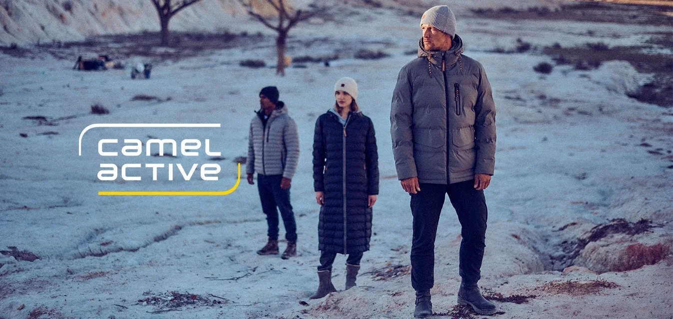 Tre persone si trovano su un paesaggio roccioso e chiaro. L'uomo davanti indossa un giubbotto grigio imbottito, mentre gli altri indossano abiti scuri con berrettini.