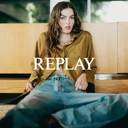 Giovane donna con una camicia verde oliva e jeans blu, seduta per terra appoggiata a un mobile di legno all'interno.