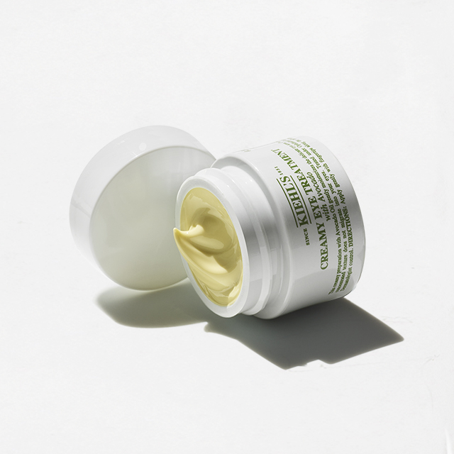 Vasetto bianco di Creamy Eye Treatment with Avocado Kiehl&rsquo;s aperto, con crema di colore verde chiaro visibile all&rsquo;interno, su sfondo chiaro.