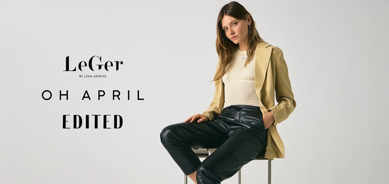 Giovane donna seduta su uno sgabello, vestita con un blazer beige, un maglione crema, pantaloni in pelle nera e sandali con tacco neri, su uno sfondo chiaro e uniforme.