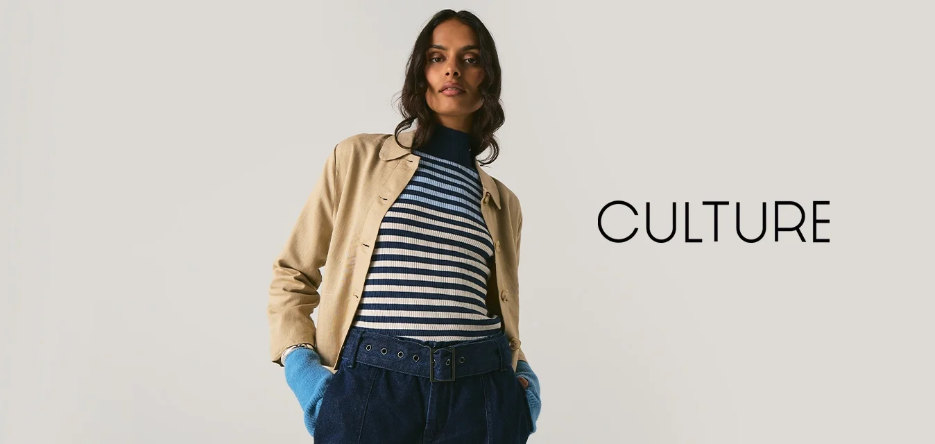 Persona che indossa un giaccone beige, un maglione a righe, una cintura larga, pantaloni scuri e guanti blu brillanti, in piedi contro uno sfondo chiaro e uniforme.