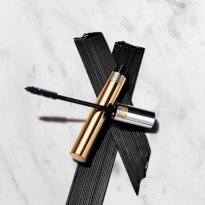 Un tubetto di mascara YSL dorato e il suo applicatore esposti su una superficie di marmo bianco.