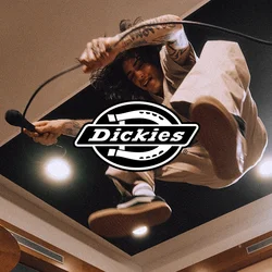 Cantante con tatuaggi salta in uno studio musicale tenendo un microfono, un batterista suona dietro un set di batteria, attrezzature moderne per studio sono visibili.